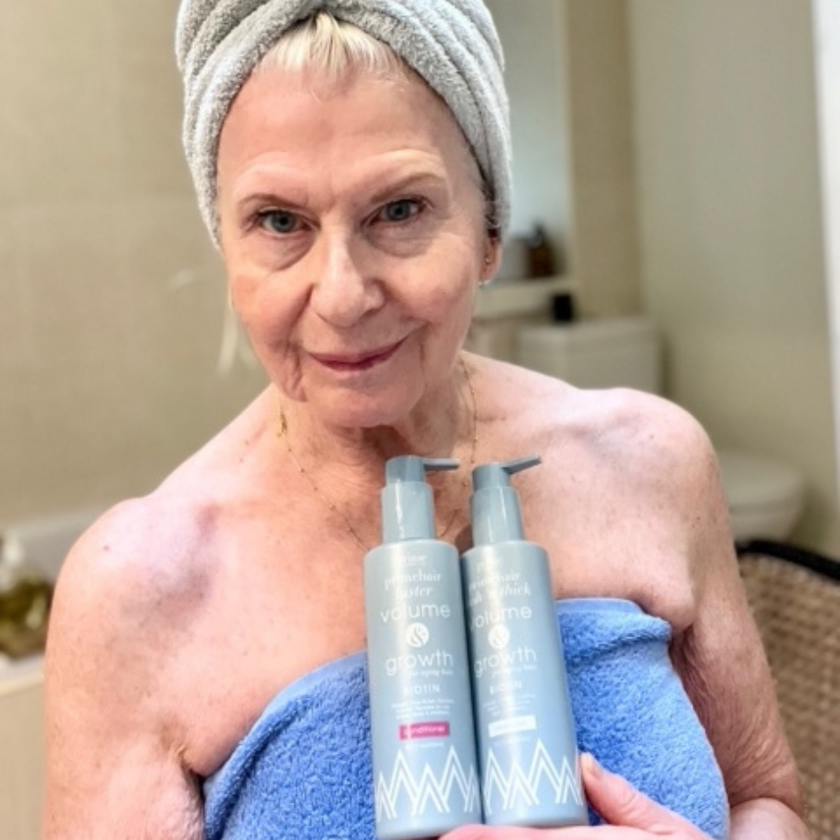 An older woman with a towel wrapped around her head and shoulders smiles while holding two bottles of hair products in a bathroom.