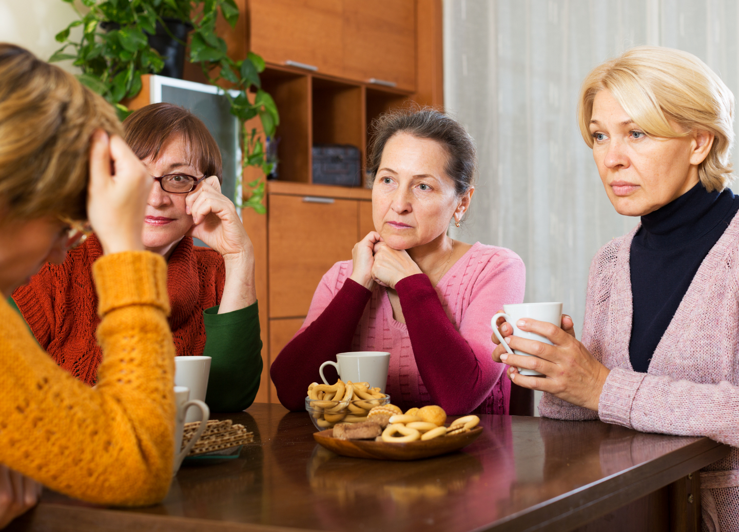 Four women sit around a table with mugs and snacks, engaged in a serious conversation. They appear thoughtful and concerned, with one woman resting her head on her hand.