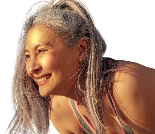 A smiling woman with long gray hair, wearing a sports tank top, looks to the side in warm sunlight against a white background.