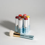 Six colorful makeup sticks with metallic blue bases from PrimePrometics™' Build Your Own Bundle are arranged in a semicircle next to a soft-bristled "Prime" makeup brush, all set against a light gray background.