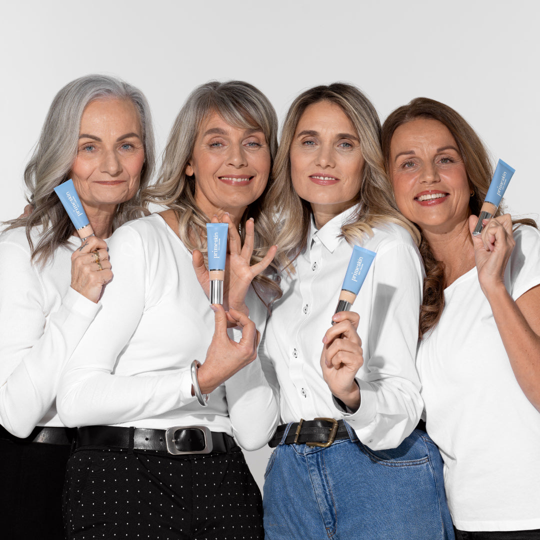 Four women with gray and light brown hair, wearing white tops, stand together smiling and holding blue skincare or cosmetic tubes against a plain light background.
