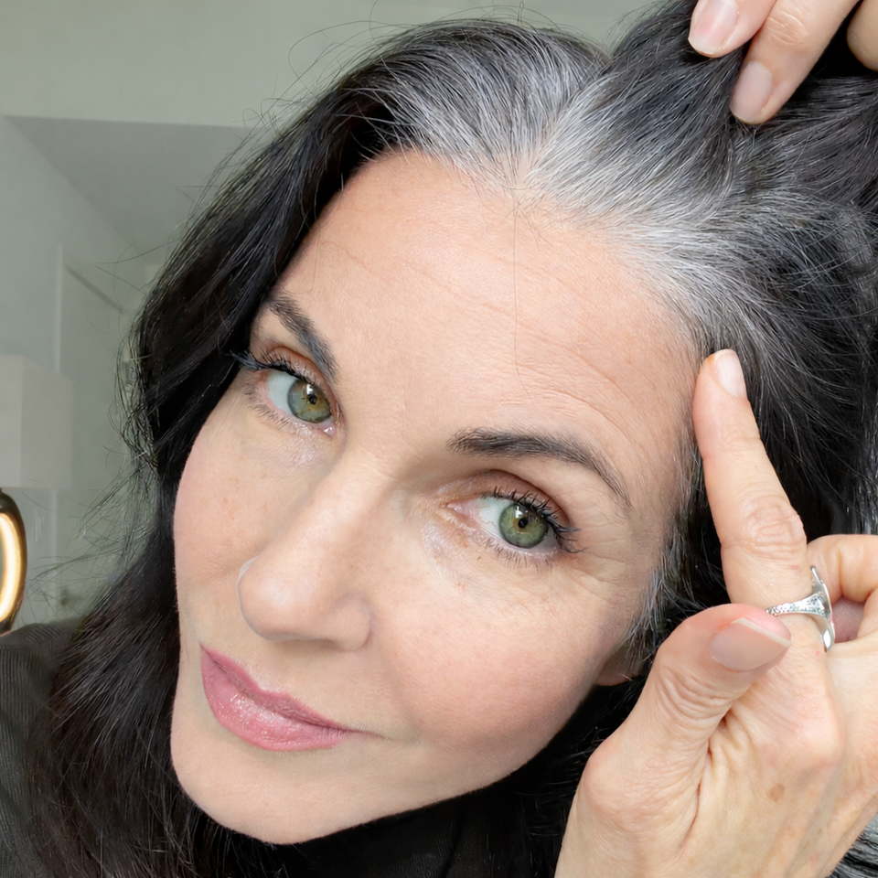 A woman with green eyes and long dark hair, streaked with gray, holds her hair to reveal her natural roots. She wears a ring and has a subtle, confident expression.