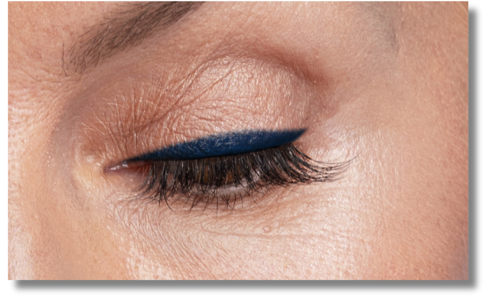A close-up of a closed eye with natural makeup, featuring bold dark blue eyeliner and long eyelashes on smooth, light skin.