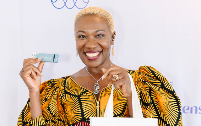A smiling woman with short blonde hair, wearing a vibrant yellow and black patterned dress, holds up a small teal-colored product box in one hand and a white bag in the other, standing in front of a white backdrop.