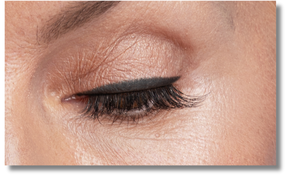 A close-up of a persons closed eye showing smooth skin, well-defined eyebrow, black eyeliner, and long curled eyelashes.