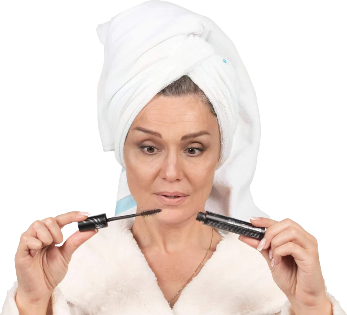 A woman with a towel wrapped around her head and wearing a bathrobe holds an open mascara wand, looking at it with a thoughtful expression, against a plain light background.