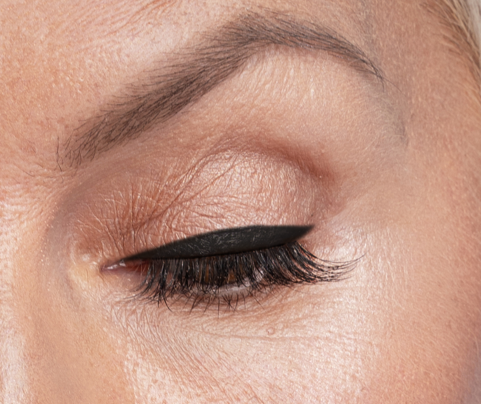 Close-up of a closed eye with neatly groomed eyebrow, smooth skin, neutral eyeshadow, and a sharp, winged black eyeliner on the upper eyelid.