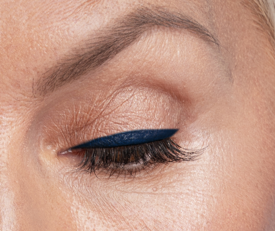 A close-up of a closed eye with smooth skin, dark blue winged eyeliner, long lashes, and a well-groomed eyebrow.