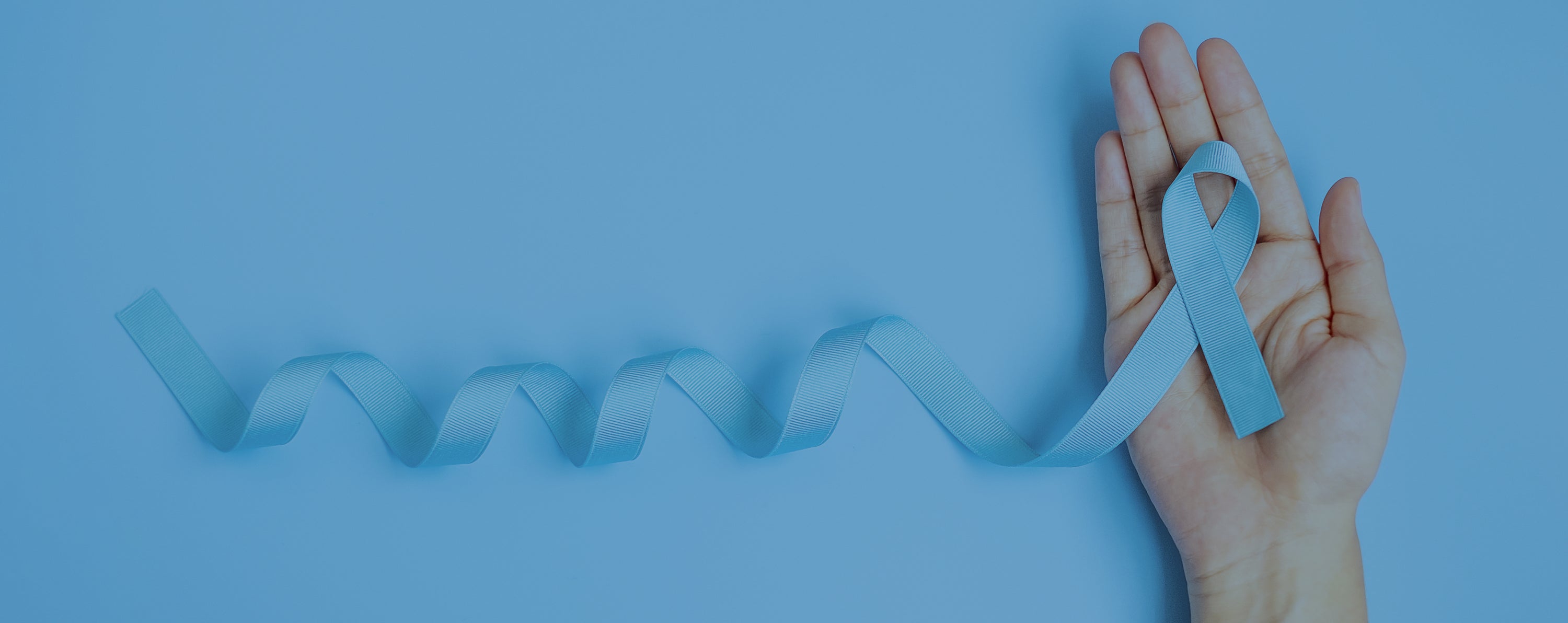 A hand holding a light blue awareness ribbon, with the ribbon extending and curling on a solid light blue background.