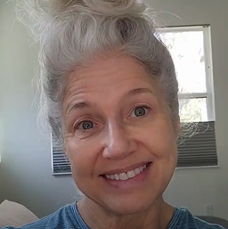 A smiling older woman with long gray hair in a messy bun, wearing a blue top, sits indoors with sunlight coming through the windows behind her.